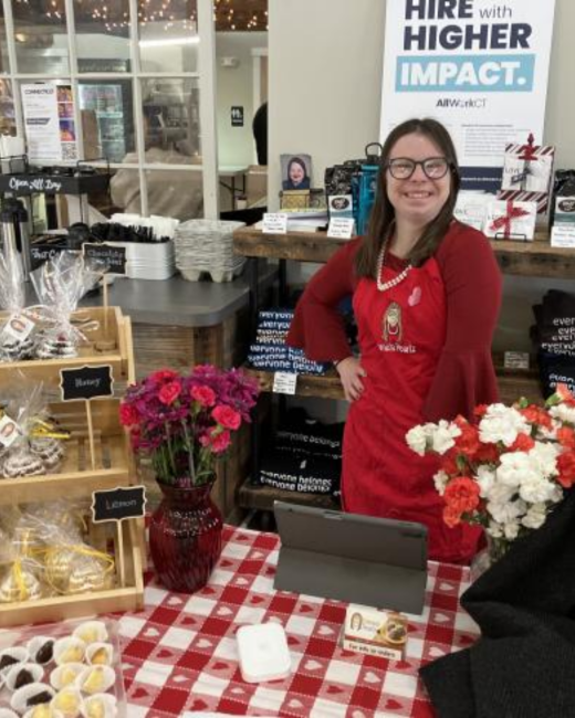 A smiling woman with Down syndrome, long dark brown hair, and glasses stands with one hand on her hip behind a table with a red-and-white checkered, heart-patterned cloth. She wears a red bell-sleeved top, a pearl necklace, and a red apron with a small logo. The table holds a vase of pink flowers, a tray of small chocolates, a bouquet of red and white carnations, and wooden crates of wrapped treats with chalkboard labels. A sign behind her reads “Hire with higher impact.” and “AllWorkCT.”