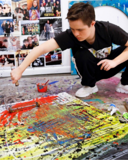 A young man with Down syndrome and short brown hair crouches on a paint-splattered floor and rolls red paint across a large abstract canvas with yellow, green, blue, and black splatters. He wears a black T-shirt with a graphic, black track pants with a white stripe, and white sneakers. A cup of red paint sits beside him, and a wall covered in photographs is behind him.