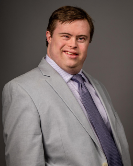 Studio headshot of a smiling man with Down syndrome and short brown hair against a dark gray background. He wears a light gray suit jacket, a lavender checked collared shirt, and a purple patterned tie.