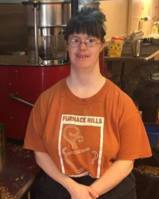 A woman with Down syndrome, dark hair in a bun and bangs, and wire-rimmed glasses sits with her hands clasped in front of a red coffee roaster and stainless steel equipment. She wears a burnt-orange T-shirt with the text “Furnace Hills” in white above a rectangular design of swirling steam lines.