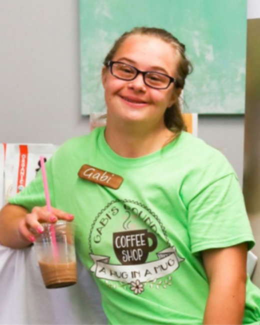 A smiling young woman with Down syndrome, brown hair pulled back, and dark-rimmed glasses holds up an iced coffee drink in a clear cup with a pink straw. She wears a lime green T-shirt with a brown name tag that reads “Gabi” and a round logo that reads “Gabi’s Grounds Coffee Shop” and “A hug in a mug,” showing a coffee mug, a ribbon banner, and a flower. A teal painting hangs on the wall behind her.
