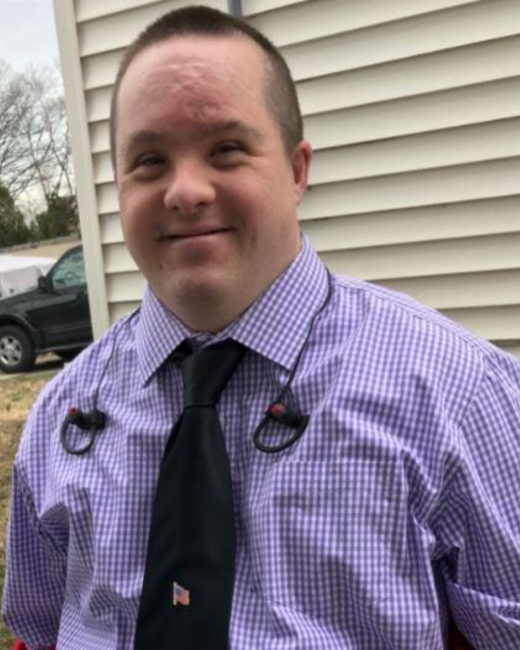 Man with Down syndrome and short dark hair smiles slightly as he stands outside next to beige house siding. He wears a purple gingham button-up shirt and a black tie with a small American flag pin, with wired earbuds hanging around his neck. A dark vehicle is parked behind him.