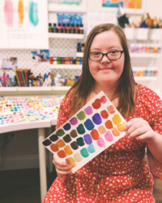 A young woman with Down syndrome and long, straight light brown hair and dark-rimmed glasses holds up a sheet of paper covered in painted color swatches in a rainbow of shades. She wears a red dress with a small white floral print and sits in an art studio with shelves of paint bottles, brushes, and more swatches on a white table behind her.