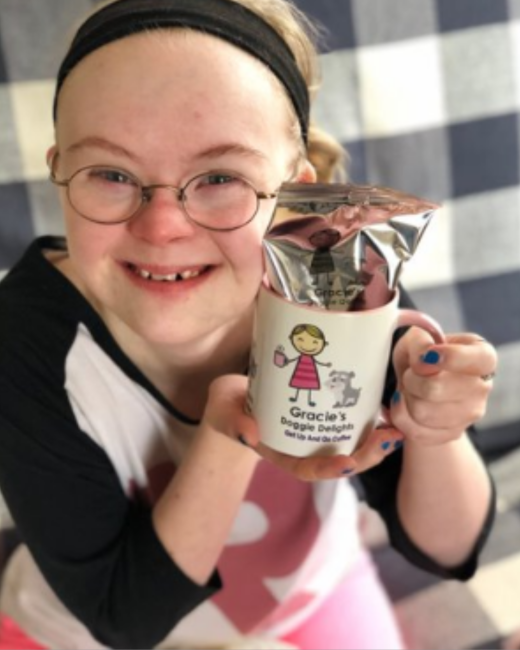 Smiling young woman with Down syndrome, blond hair, a black headband, and round wire-rimmed glasses holds up a white mug with a silver foil coffee bag inside. The mug shows a cartoon girl in a pink dress holding a cup beside a small gray dog, with the text “Gracie’s Doggie Delights” and “Get up and go coffee.” She wears a black-and-white top with a pink pattern and blue nail polish and sits against a blue-and-white checkered background.