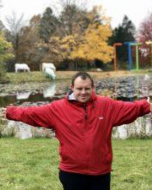 A man with Down syndrome, short dark hair, and a red zip-up jacket stands on grass with his arms stretched wide and a broad smile. Behind him is a pond with lily pads, trees in yellow and orange fall foliage, and colorful metal sculptures at the water’s edge.
