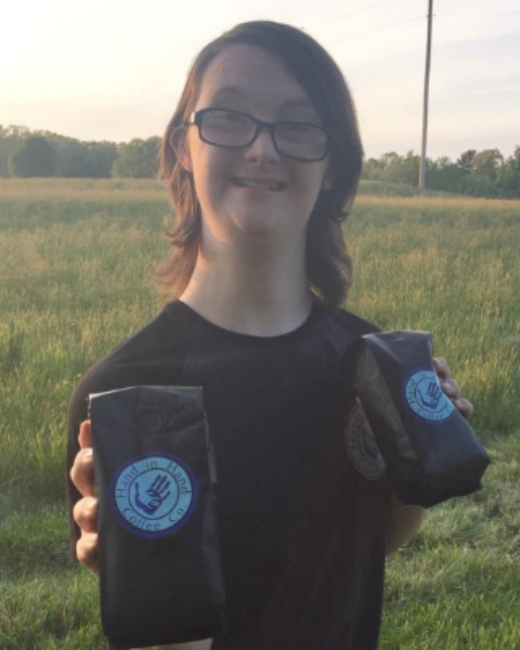 A young man with Down syndrome, shoulder-length brown hair, and dark-rimmed glasses smiles as he holds up two black bags of coffee in a grassy field at dusk. He wears a black T-shirt. Each bag has a round blue logo with a hand icon and the text “Hand in Hand Coffee Co.”