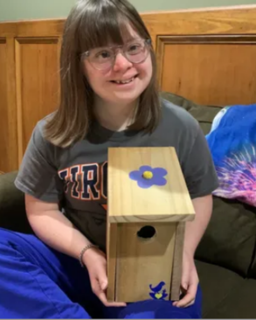 A smiling young woman with Down syndrome, straight brown hair with bangs, and pale-framed glasses sits on a dark couch holding a wooden birdhouse. The birdhouse has a purple flower on its roof and a small blue bird with a yellow heart near the base. She wears a gray T-shirt with orange-outlined lettering and a bracelet, and a blue blanket is beside her.