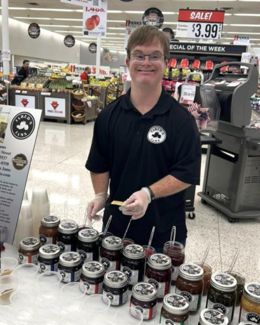 Smiling man with Down syndrome, short light brown hair, and glasses stands at a sample table in a grocery store, holding a cracker in a gloved hand. He wears a black polo shirt with a round white “Jared’s Jams” logo. Rows of jam jars with sample spoons sit in front of him, and a sign at left shows the Jared’s Jams logo and a Missouri logo.