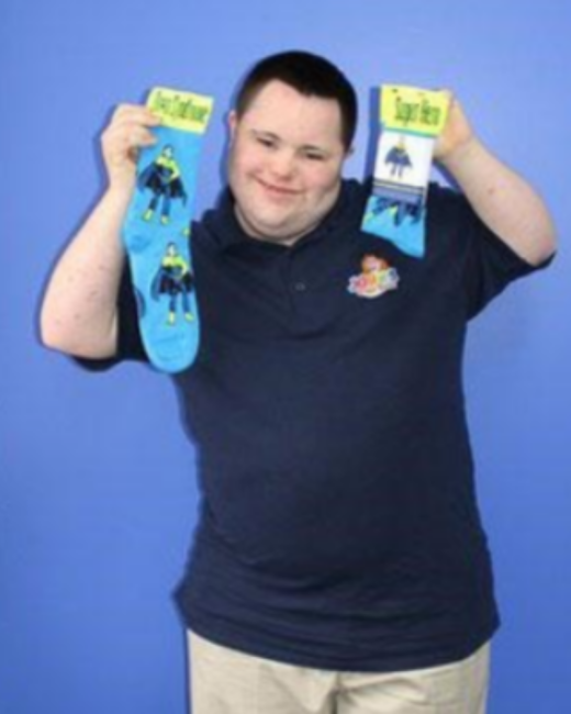 A smiling man with Down syndrome and short dark hair holds up two blue superhero-print socks, one in each hand, against a solid blue background. He wears a navy polo shirt with a small colorful logo and khaki pants.