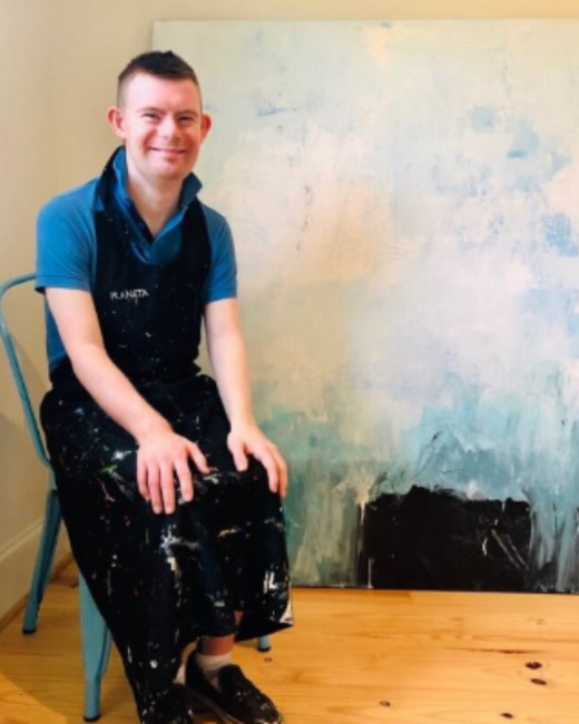 A smiling man with Down syndrome and short dark hair sits on a teal metal chair beside a large abstract painting in pale blue, white, and black. He wears a blue polo shirt, a black paint-splattered apron, black paint-splattered pants, and black shoes, and sits on a wood floor.