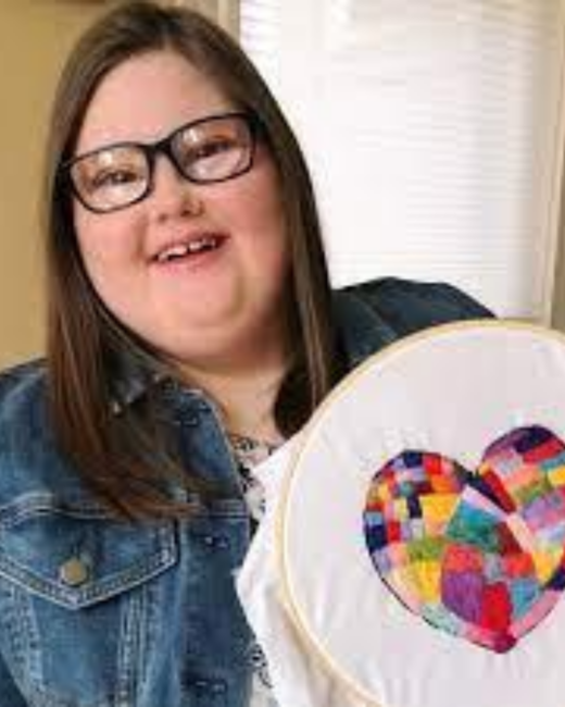 A smiling young woman with Down syndrome, long straight brown hair, and black-rimmed glasses holds up an embroidery hoop showing a heart stitched in patchwork squares of rainbow colors. She wears a denim jacket over a white patterned top.