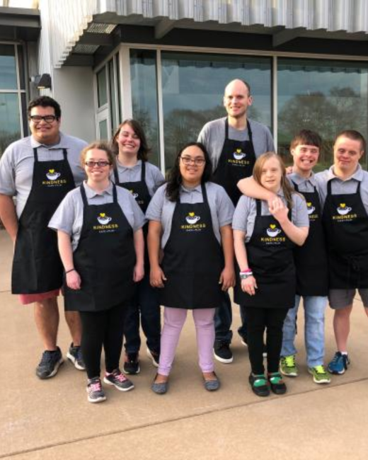 Group of eight smiling people, including individuals with Down syndrome, stand close together on a sidewalk in front of a glass building. All wear gray polo shirts and black aprons with a logo of a coffee cup with a yellow heart above the word “Kindness.”