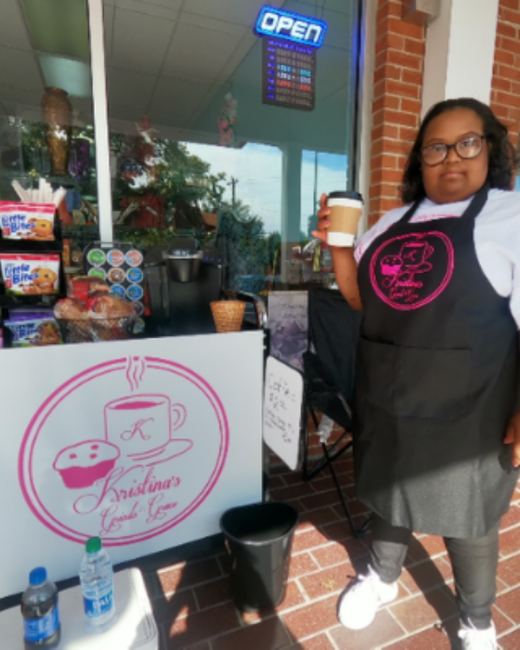 A woman with Down syndrome, dark wavy hair, and glasses stands at an outdoor coffee stand in front of a brick storefront, holding a paper coffee cup with a brown sleeve. She wears a white shirt and a black apron with a round pink logo of a coffee cup and muffin. A white sign in front of the stand shows the same logo with the script text “Kristina’s Grinds and Grace,” and snacks sit on shelves behind it.