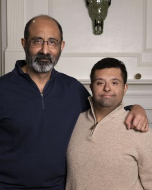 A father and his son, a man with Down syndrome, stand close together in front of a white paneled wall with a decorative animal skull above them. The man at left is bald with a gray beard and glasses, wears a navy quarter-zip sweater, and has an arm around the man at right, who has short dark hair and wears a beige quarter-zip sweater.