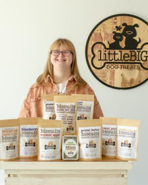 A smiling young woman with Down syndrome, long strawberry-blond hair, and glasses stands behind a wooden crate stacked with kraft paper pouches of dog biscuits in flavors including blueberry, peanut butter, sweet potato, pumpkin and peanut butter, and organic apple, plus a tin of mints. She wears a tan jacket over a white shirt. Behind her hangs a round wooden sign with a silhouette of a large and a small dog above a bone shape with the text “littleBIG” and “Dog treats.”