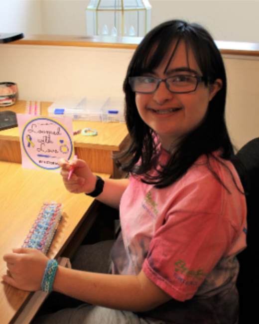 A smiling young woman with Down syndrome, long black hair with bangs, and dark-rimmed glasses holds a pink loom hook at a wooden desk. She wears a pink tie-dye T-shirt, a black watch, and a blue woven bracelet. A loom with blue and pink rubber bands lies on the desk in front of her, and a pink sign behind it reads “Loomed with Love” with a yellow and blue awareness ribbon.