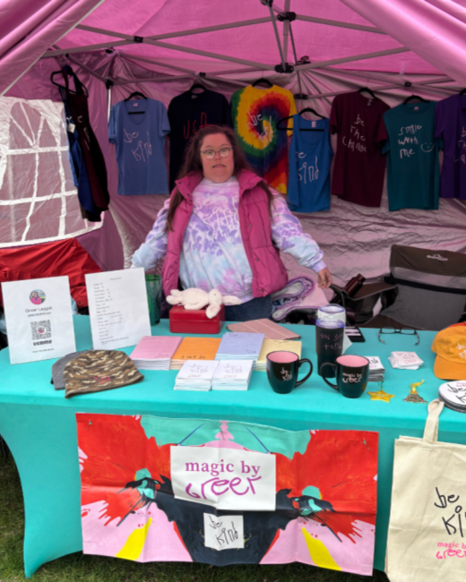 A woman with Down syndrome, long brown hair in pigtails, and glasses sits in a booth under a pink canopy. She wears a pink puffer vest over a lavender tie-dye long-sleeved shirt. T-shirts in blue, black, rainbow tie-dye, maroon, teal, and purple hang behind her with hand-lettered messages such as “Be kind,” “Be the change,” and “Smile with me.” The teal tablecloth holds notepads, mugs, hats, a stuffed bunny, and a sign with a QR code. A paint-splatter banner on the front of the table reads “Magic by Greer”