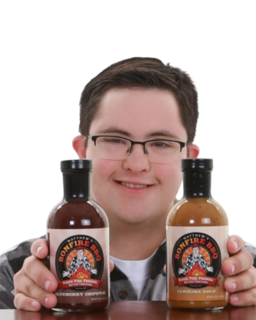 A smiling young man with Down syndrome, short dark brown hair, and dark-rimmed glasses holds two bottles of barbecue sauce in front of him against a white background. He wears a gray plaid shirt over a white T-shirt. The round labels show a cartoon man in front of a fire with the text “Matthew’s Bonfire BBQ” and the slogan “Food. Fire. Friends. Better together.” The flavors are “Blueberry chipotle” and “Carolina gold.”
