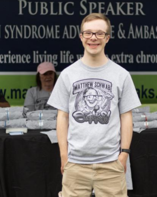 A smiling young man with Down syndrome, short light brown hair, and dark-rimmed glasses stands with his hands in the pockets of his khaki shorts. He wears a gray T-shirt with a cartoon of a man holding a microphone and the text “Matthew Schwab Speaks!” and a black watch. Behind him, a black banner reads “Public Speaker” along with partly visible lines about a Down syndrome advocate and ambassador, and a woman in a pink cap sits at a table with stacks of gray shirts.