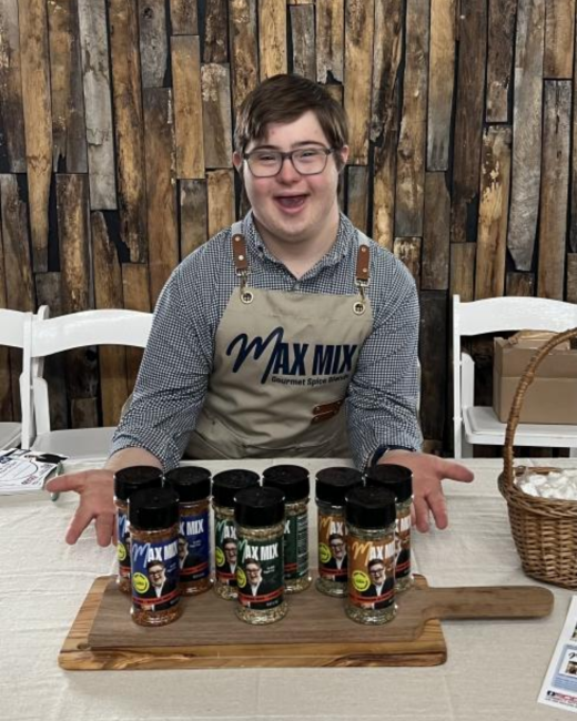 A smiling young man with Down syndrome, short brown hair, and glasses holds his hands out toward a row of eight spice shakers with black lids on a wooden board. He wears a blue-and-white gingham shirt and a tan apron with the logo “Max Mix Gourmet spice blends.” Behind him are a rustic wooden plank wall, white chairs, and a wicker basket.