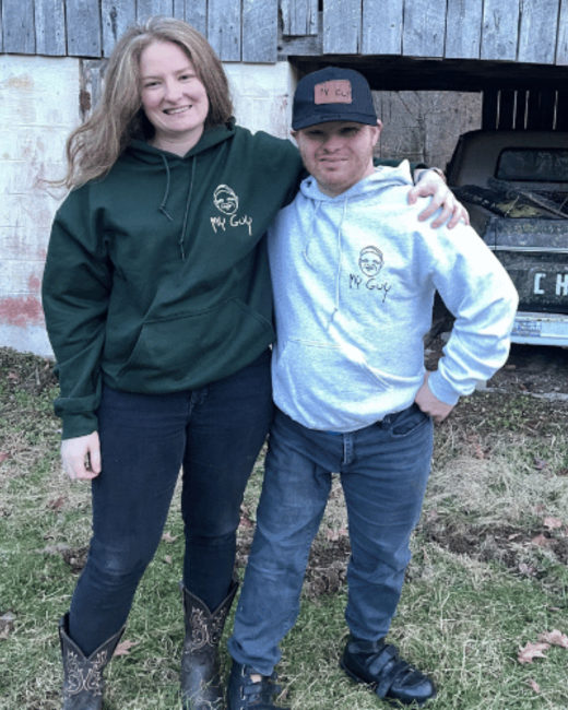 Two people stand with their arms around each other on grass in front of a weathered wooden barn and an old truck. At left, a smiling woman with long, wavy light brown hair wears a dark green hoodie with a small line-drawn face logo and the text “My guy,” along with jeans and cowboy boots. At right, a man with Down syndrome wears a navy cap with a leather patch reading “My guy,” a light gray hoodie with the same logo, jeans, and black shoes.