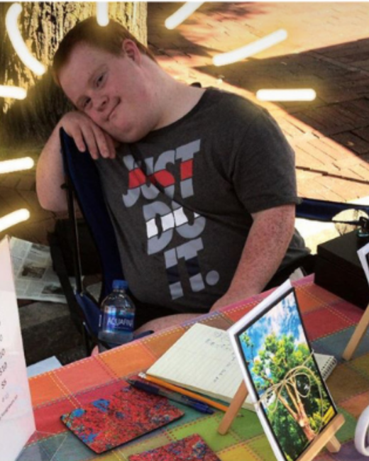 A man with Down syndrome and short red-brown hair rests his head on his hand and smiles slightly while sitting in a folding chair at a vendor table. He wears a gray T-shirt with the words “Just do it.” in white and red. The table has a bright plaid cloth and holds a notebook, pens, a water bottle, colorful coasters, and a framed photo of a green tree, with string lights and a brick pavement behind him.