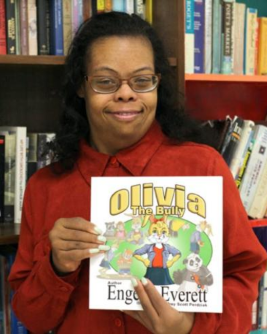 A woman with Down syndrome, with long dark curly hair, and brown-framed glasses smiles slightly while holding up a children’s book in front of bookshelves. She wears a rust-red collared shirt. The cover shows the title “Olivia the Bully” in yellow letters above an illustration of a cat-like cartoon character in a blue jacket surrounded by other animals, with the word “Author” and the name “Everett” visible below.