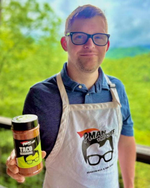 A man with Down syndrome, short brown hair, light stubble, and black-rimmed glasses smiles slightly while holding a jar of taco seasoning on a deck with green, forested mountains behind him. He wears a navy polo shirt and a white apron with a logo of a cartoon face with glasses and flame-shaped hair and the text “Pman the Chef” and “Seasonings & BBQ rubs.” The jar shows the same logo and the words “Taco seasoning.”