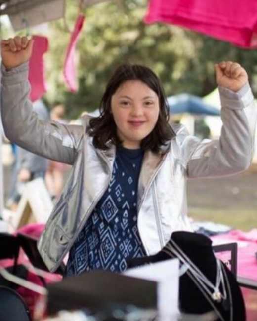 Young woman with Down syndrome and shoulder-length dark hair smiles and raises both fists overhead at an outdoor vendor booth. She wears a metallic silver jacket over a navy top with a geometric pattern. Pink items hang behind her, a jewelry display sits in the foreground, and blurred festival tents are in the background.