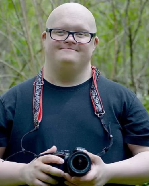 A man with Down syndrome, a shaved head, and dark-rimmed glasses smiles slightly as he holds a Nikon camera in both hands outdoors. He wears a navy T-shirt and a red-and-black patterned camera strap, with green branches and foliage behind him.