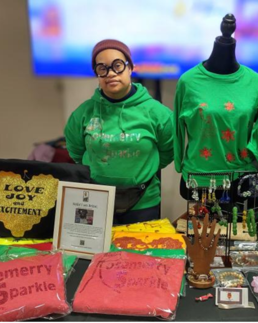 A woman with Down syndrome, a maroon knit beanie, and round black glasses stands behind a vendor table in a green hoodie with glittery lettering that reads “Rosemerry Sparkle.” Beside her, a dress form wears a green sweatshirt with red star designs. The table holds red shirts in plastic bags printed with “Rosemerry Sparkle,” yellow and orange shirts with the word “Love,” jewelry displays, a hand-shaped bracelet stand, and a black bag with a gold glitter heart reading “Love joy and excitement.”