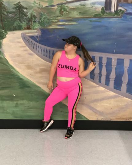 A young woman with Down syndrome and a long ponytail leans against a painted mural of a lake and stone balustrade, holding the end of her ponytail with one hand. She wears a black sparkly baseball cap, a hot pink crop tank top that reads “Zumba,” matching hot pink leggings with black and gold side stripes, and black and gold sneakers.