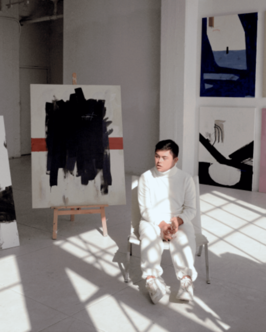 A young man with Down syndrome and short dark hair sits on a white chair in a sunlit white studio, looking off to the side with his hands clasped. He wears a white turtleneck sweater, white pants, and white sneakers. Large abstract paintings in black, navy, white, and red surround him, including one on an easel with thick black brushstrokes and a red stripe.