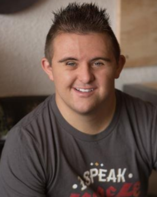 A man with Down syndrome and short, spiky brown hair smiles widely at the camera. He wears a gray T-shirt with the words “I speak” in white above red lettering that is cut off at the bottom of the frame. The background is softly blurred.