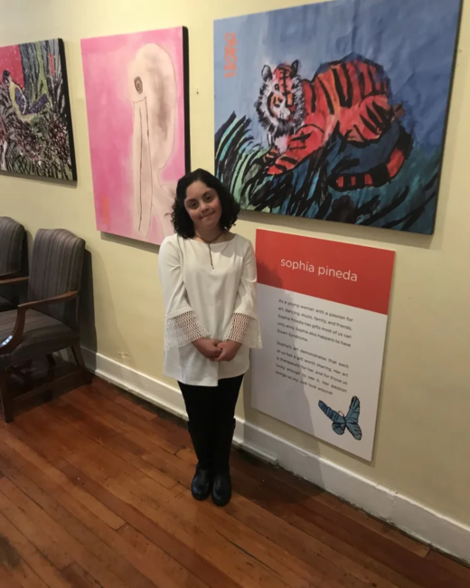A woman with Down syndrome and short, dark curly hair stands with her hands clasped in a gallery on a wood floor. She wears a white long-sleeved blouse with lace cuffs, black pants, and black boots. Beside her is a wall sign with an orange header reading “sophia pineda,” a paragraph about her art, and a blue butterfly illustration. Above her hang three paintings: an orange and black tiger on a blue background, an abstract figure on pink, and a dark floral scene.