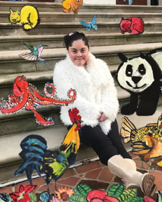 A young woman with Down syndrome and dark hair pinned up with a small clip smiles slightly as she sits on stone steps. She wears a white fluffy coat, black pants, and cream boots. Colorful painted animals and flowers surround her, including a yellow cat, a red cat, a blue butterfly, a hummingbird, a red octopus, a panda, a rooster, a hen, and red flowers with green leaves.