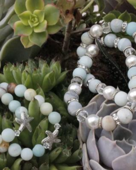 Close-up of beaded bracelets in pale blue stones, white pearls, and clear crystals with silver spacers and silver starfish charms, resting on green and gray-lavender succulent plants.