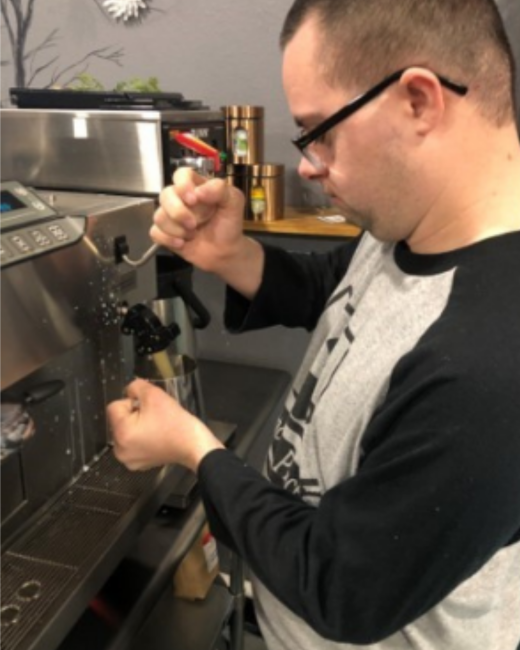 A man with Down syndrome, short dark hair, and black-framed glasses is shown in profile as he looks down and steams milk in a metal pitcher at a stainless steel espresso machine. He wears a gray and black baseball-style shirt with black sleeves. Copper canisters sit on a shelf behind him.