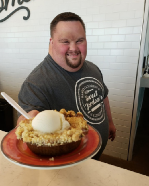 A smiling man with Down syndrome, short dark hair, and a goatee holds out a red plate with a crumble dessert topped with a scoop of vanilla ice cream and a white spoon. He wears a gray T-shirt with a round logo that reads “Baked goodies & Sweet Jordan’s Ice cream,” with a whisk and a rolling pin. A white subway-tile wall is behind him.
