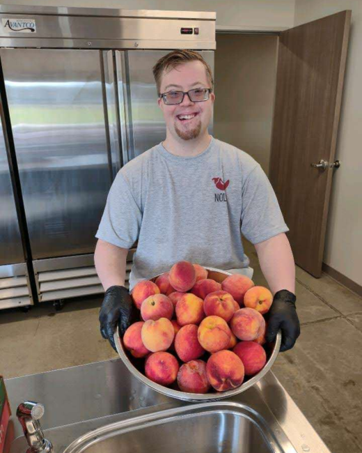 A man with Down syndrome wears a gray shirt with a logo with a red pepper, glasses, and black kitchen gloves. He stands in an industrial kitchen holding a large bowl of peaches