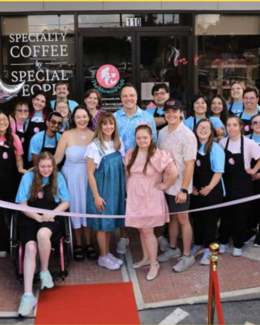 Large group of smiling people, including individuals with Down syndrome, gather outside a café storefront for a ribbon-cutting. A pink ribbon stretches across the front, with a red carpet and gold stanchion on the sidewalk. Many wear light blue shirts and black aprons, and a young woman in a wheelchair sits at front left. The window reads “Specialty coffee by special people,” cropped at the edge, beside the number 110 and a round pink mermaid logo with the words “The Mermaid Café.”