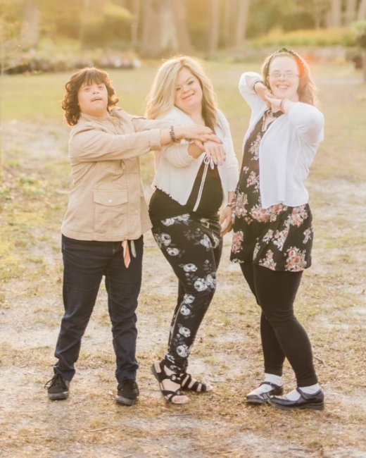 Three young women with Down syndrome pose outdoors in a sunlit field with their arms crossed together at the center. At left, a woman with dark curly hair wears a tan jacket, navy pants, and black sneakers. In the middle, a woman with long blond hair wears a white cardigan, a black top, black floral pants, and black sandals. At right, a woman with long auburn hair, a headband, and glasses wears a white cardigan, a black floral dress, black leggings, and black Mary Jane shoes with white socks.