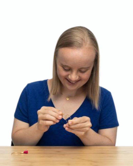 A young woman with Down syndrome and straight, shoulder-length blond hair smiles as she looks down and threads a small gold hoop earring at a light wood table. She wears a navy V-neck T-shirt and a thin gold necklace. A second earring with a pink tassel lies on the table in front of her, and the background is white.