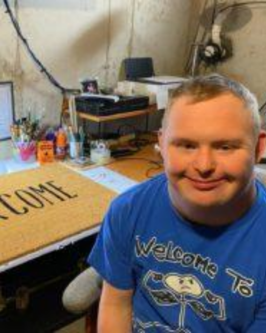 A smiling man with Down syndrome and short gray-brown hair sits at a cluttered basement desk with art supplies, a fan, and a “Welcome” doormat on top. He wears a blue T-shirt with the words “Welcome to” above a hand-drawn figure.