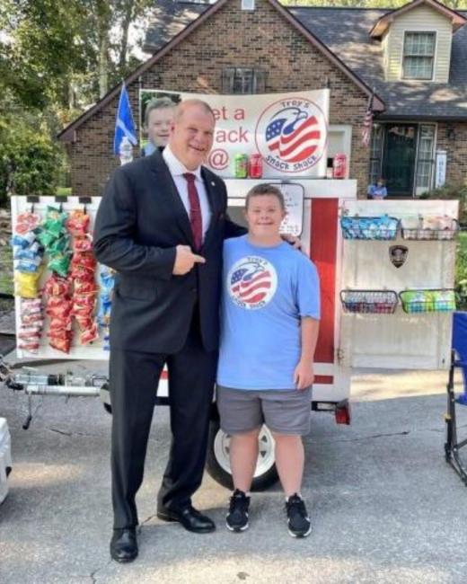 Tall man in a dark suit, white shirt, and maroon tie smiles with his arm around a boy with Down syndrome in front of a snack cart. The boy wears a light blue T-shirt with a round “Trey’s Snack Shack” logo showing an eagle with flag stripes, gray shorts, and black sneakers. Chip bags hang on a pegboard on the cart, baskets of snacks line its side, and a banner above shows the same round logo and a photo of a boy. A brick house is behind them.