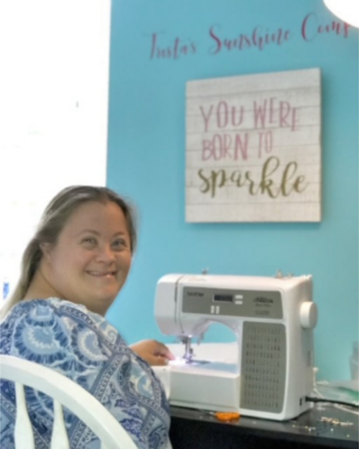A smiling woman with Down syndrome and shoulder-length light brown hair pulled back looks over her shoulder at the camera while sitting at a white sewing machine. She wears a blue-and-white paisley top. A teal wall behind her has cropped script lettering that begins “Trista’s Sunshine” and a wooden sign that reads “You were born to sparkle.”