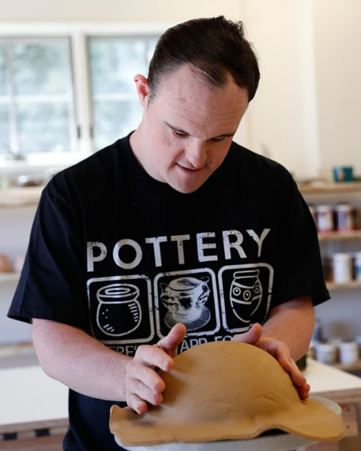 A man with Down syndrome and short dark hair looks down with concentration as he presses his hands on a flat slab of tan clay in a pottery studio. He wears a black T-shirt that reads “Pottery” above three icons of clay pots. Shelves of glazes sit behind him.
