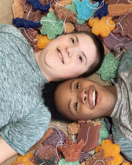 Overhead view of two smiling young men lying side by side with their heads together, surrounded by glittery flower, tulip, and teddy-shaped ornaments in orange, green, blue, purple, and brown, each with white string. The man at top has a buzzed haircut and wears a heathered green-gray T-shirt. The man at right has dark curly hair and wears a heathered gray shirt.