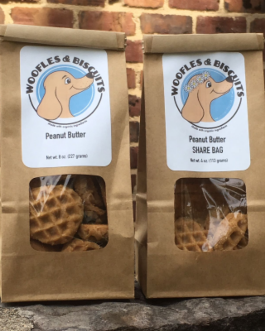 Two brown paper bags of dog treats sit on a stone ledge in front of a brick wall. Clear windows show waffle-shaped peanut butter treats. White labels show a round logo of a cartoon tan dog on a blue circle, with the text “Woofles & Biscuits” and “Made with organic ingredients.” The left label reads “Peanut butter” and “Net wt. 8 oz. (227 grams).” The right label reads “Peanut butter,” “Share bag,” and “Net wt. 4 oz. (113 grams),” and its dog wears a daisy crown.