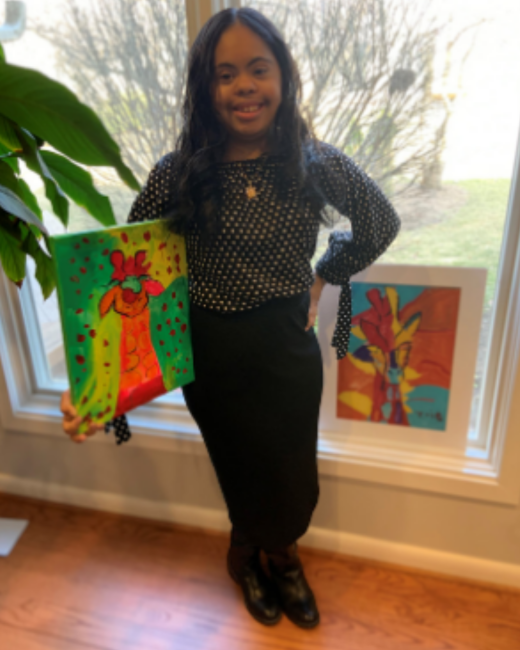 A smiling young woman with Down syndrome and long, wavy dark hair stands with one hand on her hip beside a window, holding a canvas painting of a bird in green, yellow, and red with red dots. She wears a black polka-dot blouse with tied sleeves, a black skirt, black boots, and a gold necklace. Another painting in yellow, red, and teal sits on the windowsill, and a leafy plant and wood floor are visible.